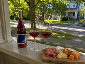 A glass of itä carbonic zinfandel on a porch windowsill next to a charcuterie board with prosciutto, cheese, crackers, and melon.