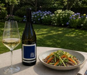 A bottle and glass of itä sémillon on an outdoor table beside a bowl of cold soba noodle salad with julienned carrots, cucumber, and scallions.